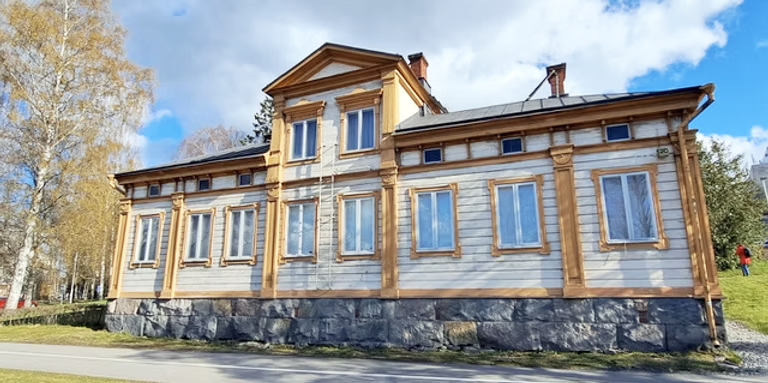 Vanha puurakennus, jolla korkea kivijalka ja keltamullatut koristelaudat.